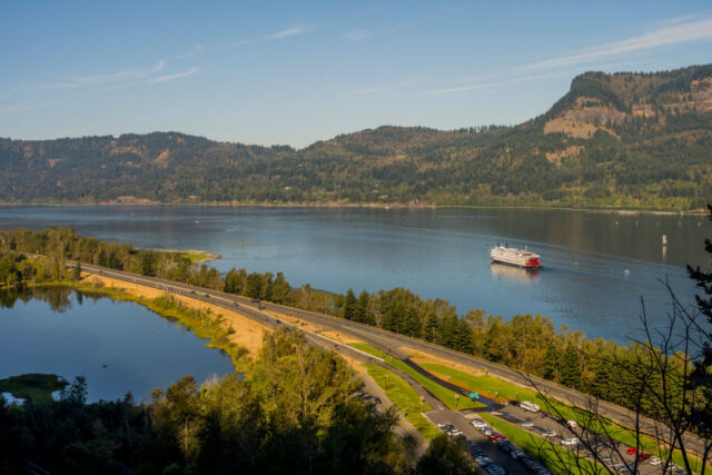 Ship sailing down the Columbia River with a bridge in the foreground and treed mountains in the background.