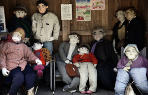 Numerous life-sized dolls, standing and sitting, in a wooden shelter.