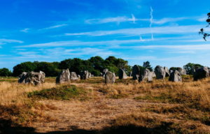 View looking out at standing stones in France across a field of dried grass with a blue sky.