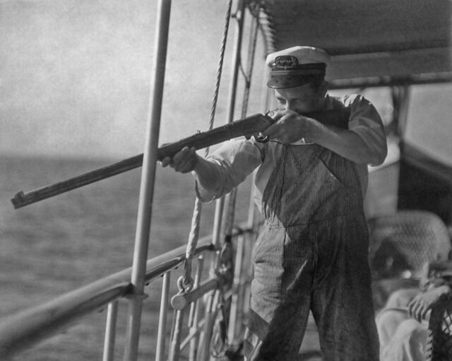 Jacob Martin aiming a rifle while standing on the deck of a ship