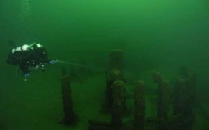 A scuba diver in murky green water examines stone ruins in a lake.