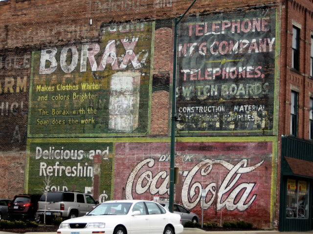 Cars parked in front of various ghost signs, which are on the exterior of a brick building